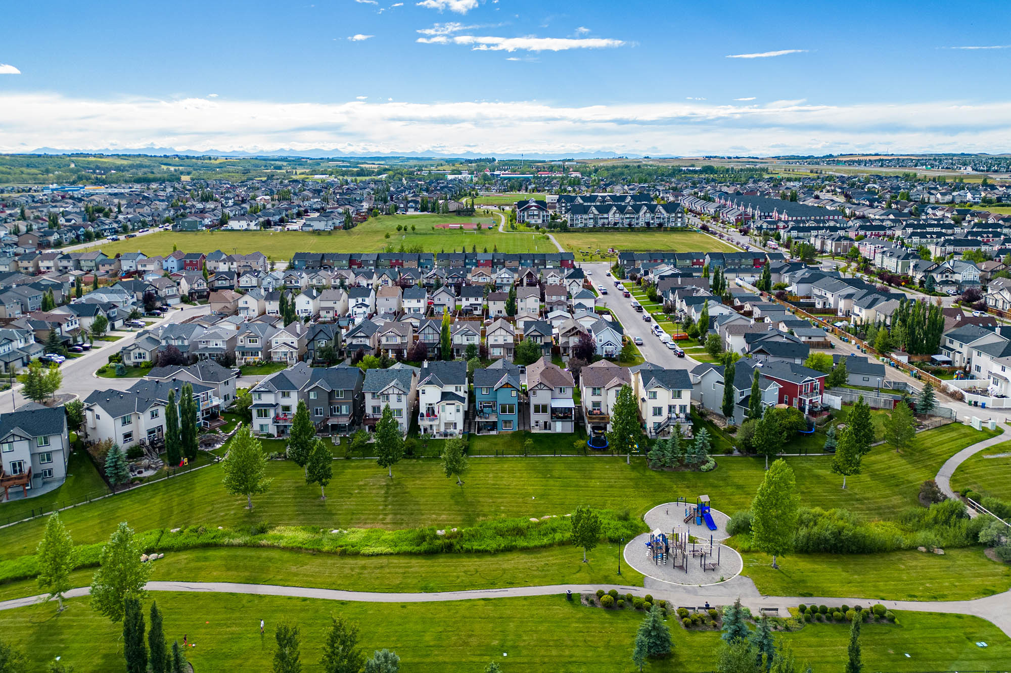 Aerial drone photograph of the Calgary Silverado community showing a playground, green space, detached homes for sale, a Silverado baseball diamond, Turner Field - a sports field, and the Rocky Mountains in the distance.