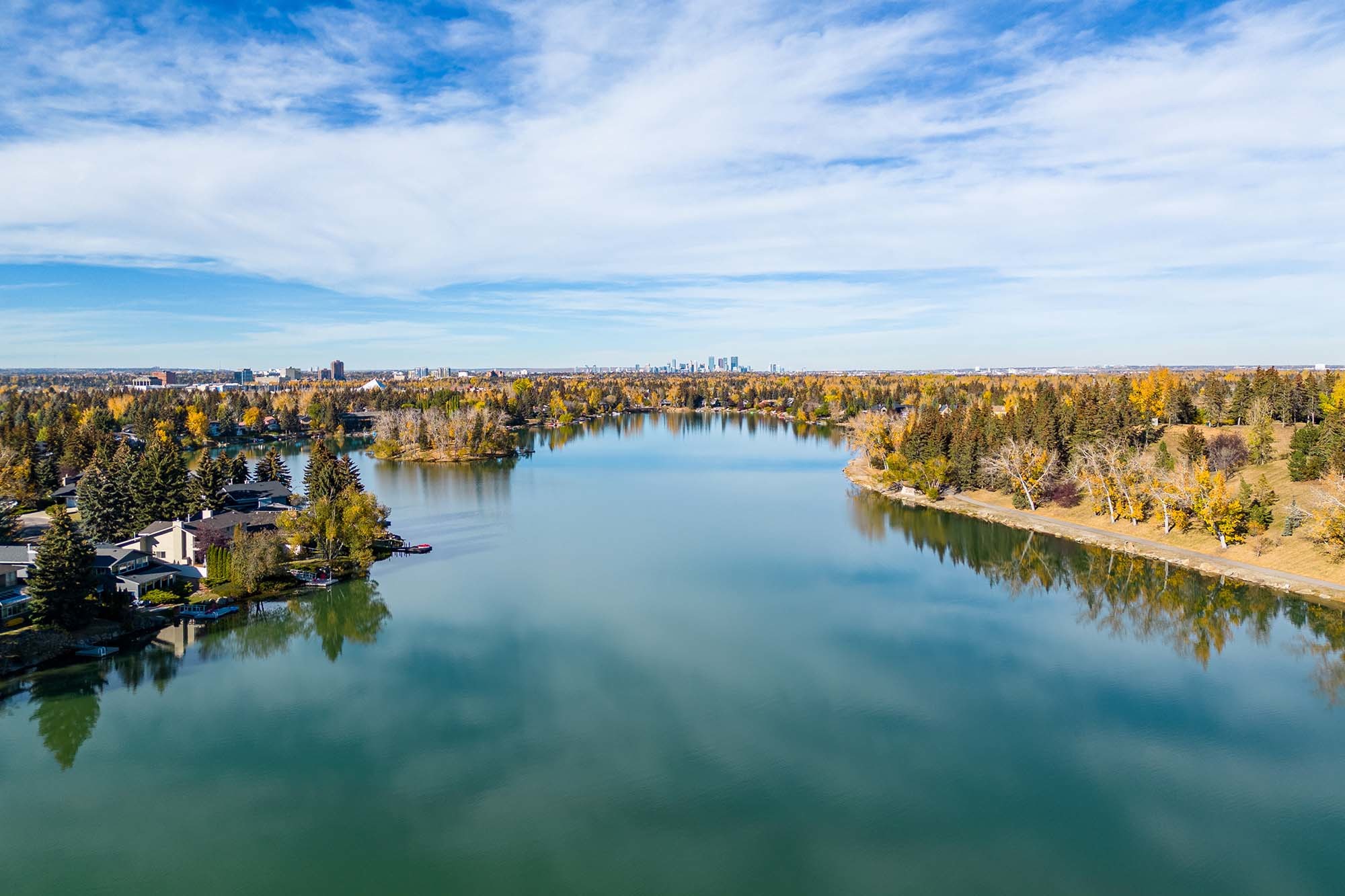 A view overlooking the water of Lake Bonavista, Calgary, Alberta, Canada with waterfront lake homes for sale and the Downtown Calgary skyline in the background.