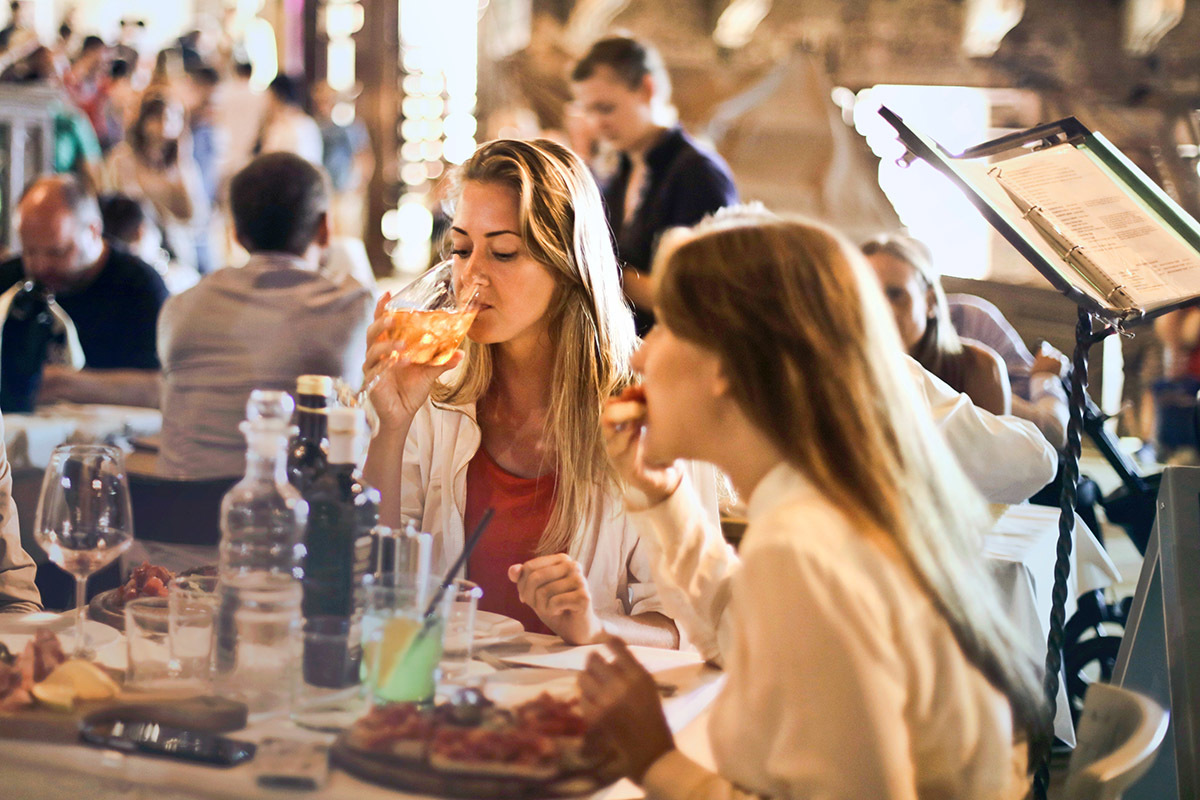 Two women eating dinner and drinking wine at a busy restaurant with people socializing in the background